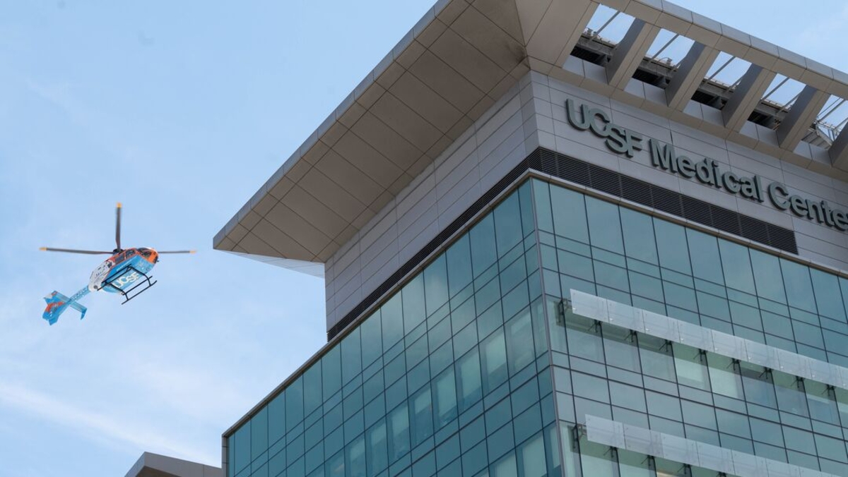 A UCSF Medical Center helicopter flies beside the corner of a modern glass hospital building, with the UCSF Medical Center sign visible near the rooftop helipad against a clear blue sky.