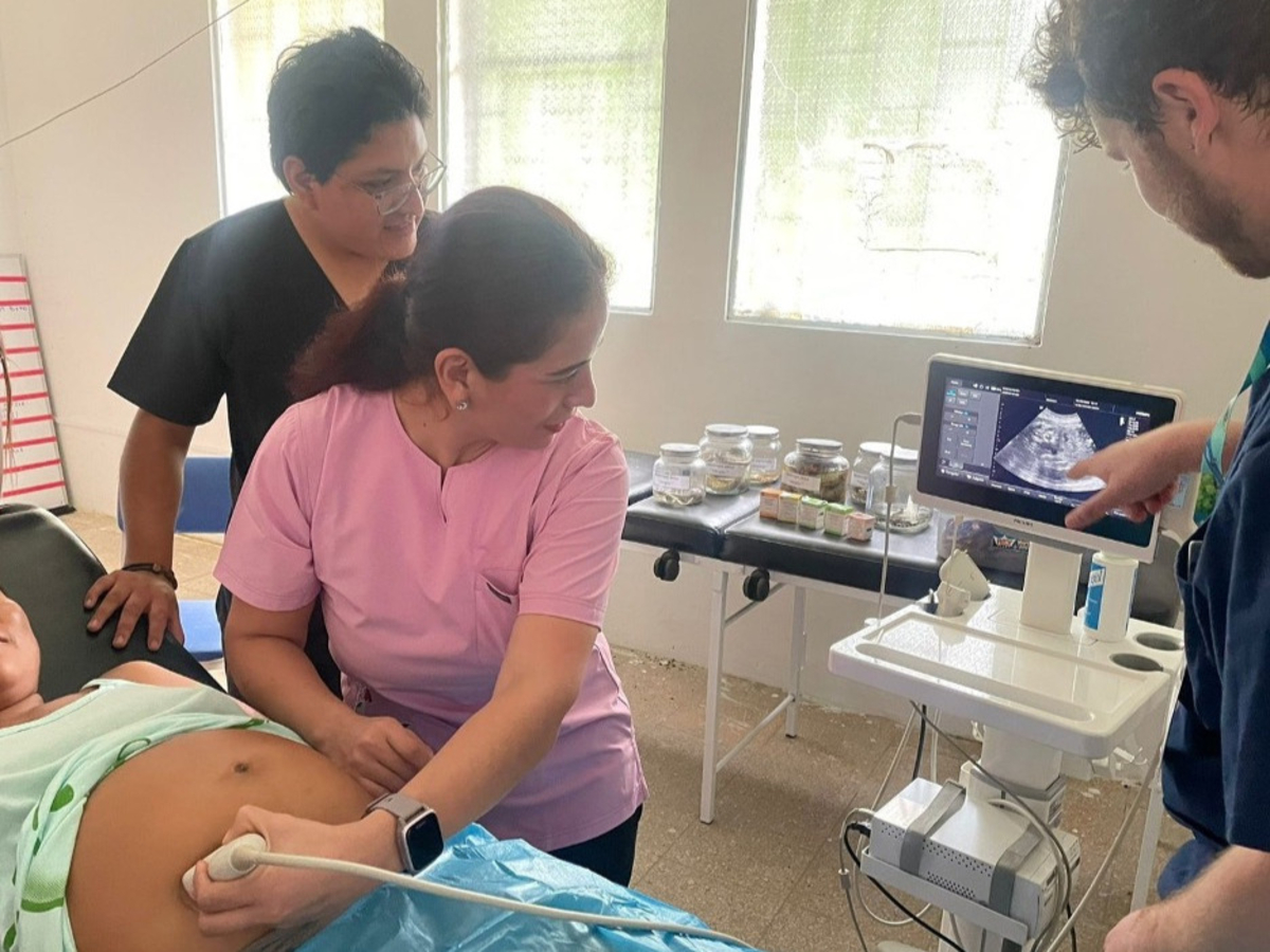 Healthcare providers participate in hands-on obstetric ultrasound training. A clinician scans a pregnant patient’s abdomen while colleagues review the ultrasound image on a nearby monitor, demonstrating point-of-care ultrasound instruction in a clinical setting.
