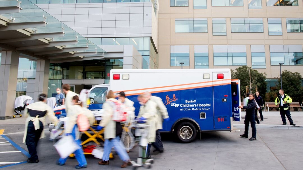 Emergency department staff transport a pediatric patient on a stretcher outside UCSF Benioff Children's Hospital at UCSF Medical Center, moving past a parked ambulance at the hospital entrance.
